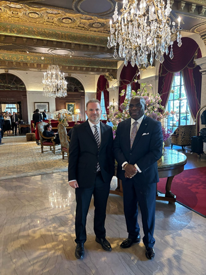 2 men standing together in a reception hall under a chandelier 