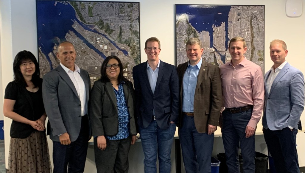 seven people in professional dress standing facing the camera in front of maps of the Port of Tacoma