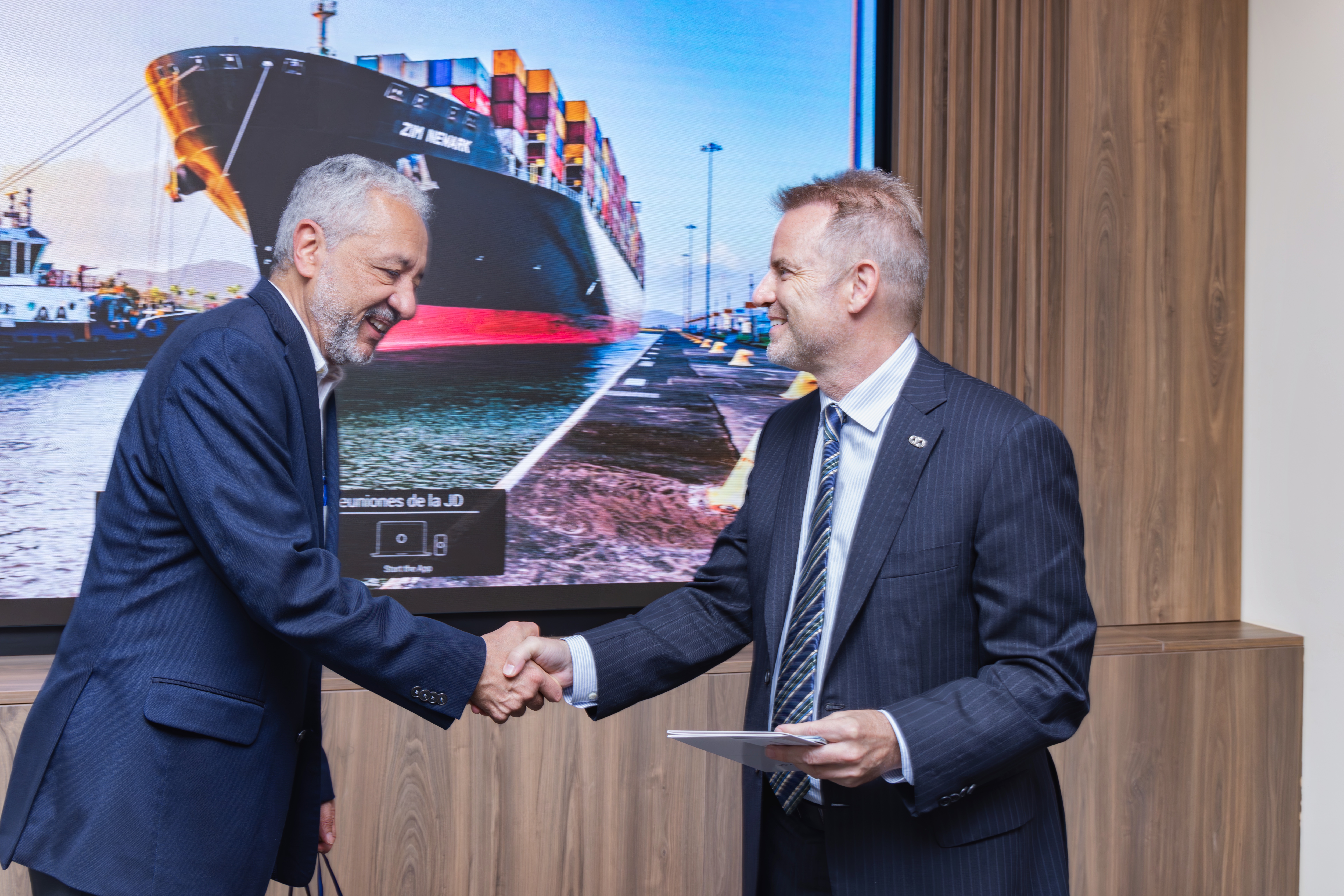 2 men in suits shaking hands in front of a large photo of a container ship