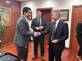 2 men in suits standing and shaking hands while a third man stands and observes