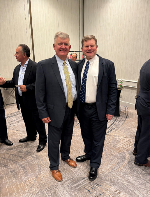 Two men in suits in a conference hall. The men are smiling into the camera.