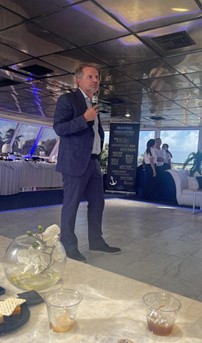 Man standing in a suit with a microphone standing in the center of the room in front of tables.