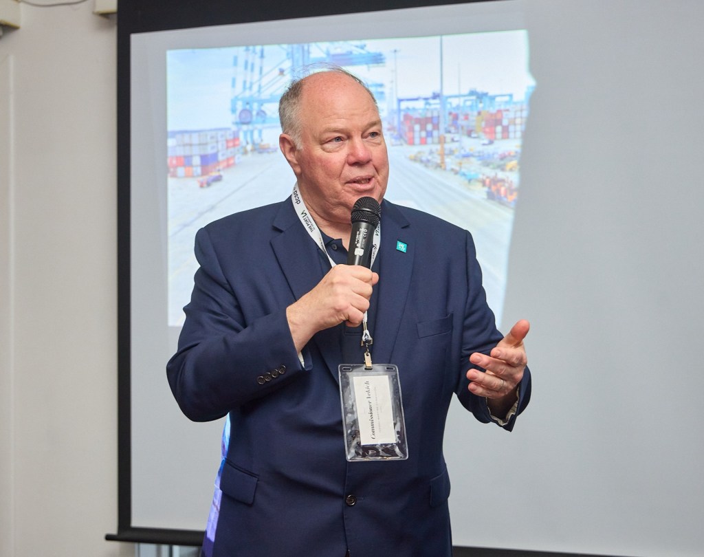 A man in a suit holding a microphone and speaking to people out of frame of the photo. The man in standing in front of a projector screen that shows many shipping containers.