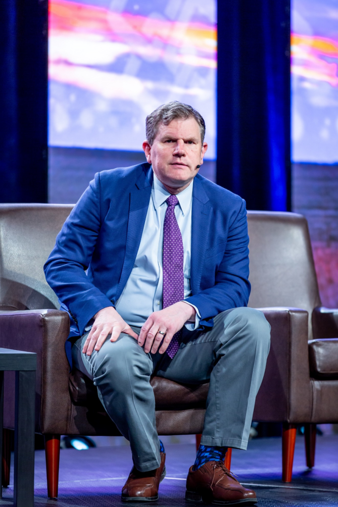 Man in a suit sitting in a chair with a mic on his face speaking.