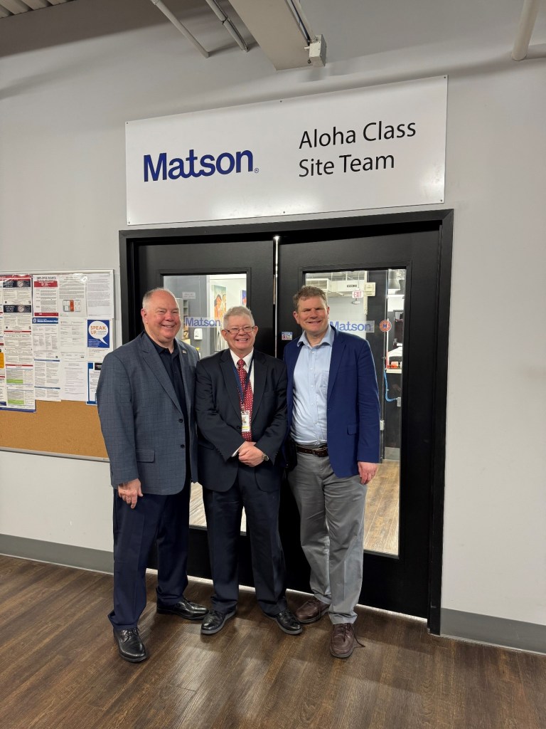 Three men standing in front of double black doors, posing for a photo under a sign that says Matson, Aloha Class Site Team