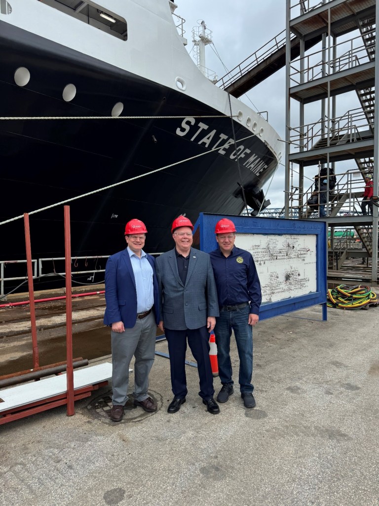 Three men wearing red hard hats, standing in front a ship that says State of Maine, posing for a photo.