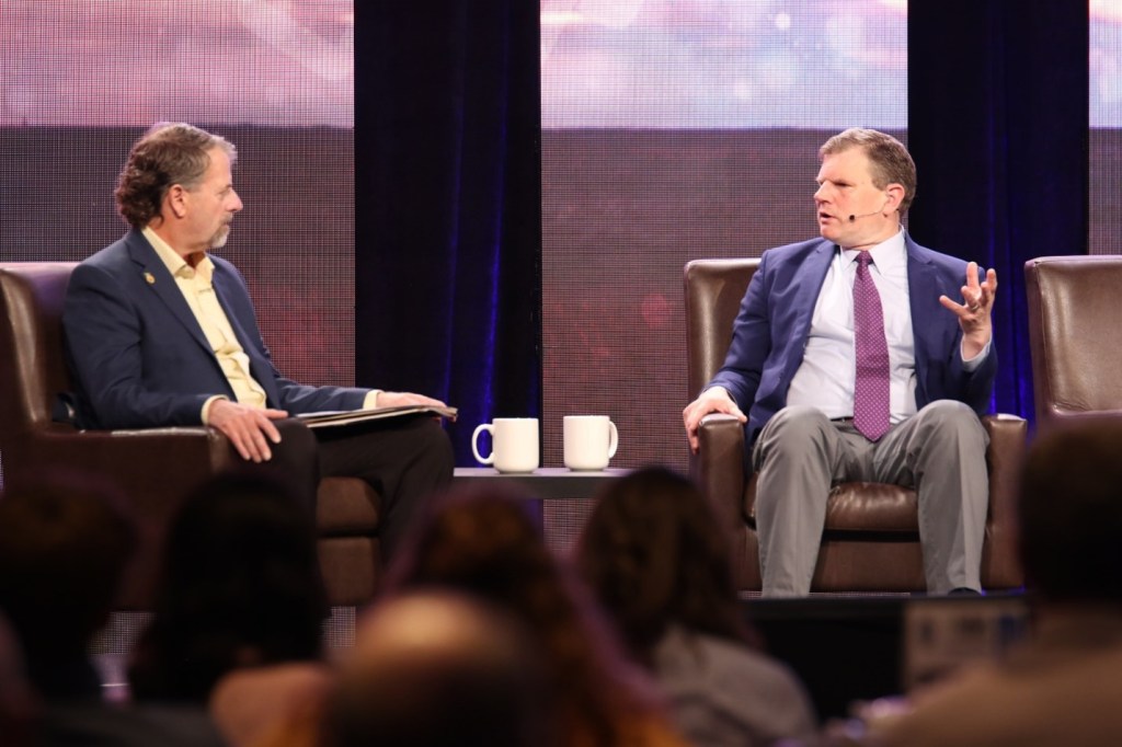 Two men sitting in chairs in front of an audience speaking to one another.