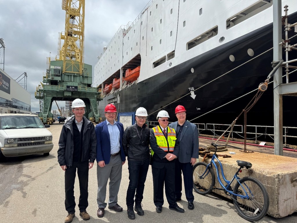 Five men with hard hats pose for a picture in front of a big ship.