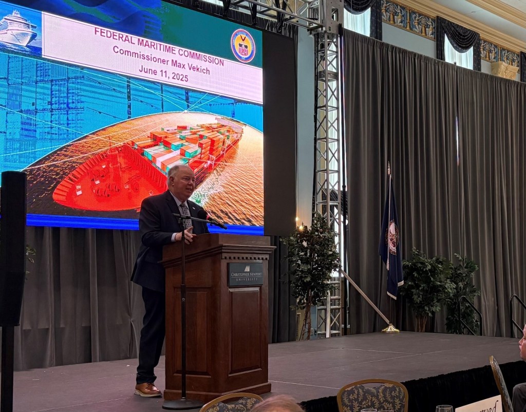 Man in suit on stage speaking in front of an image of a ship carrying containers.