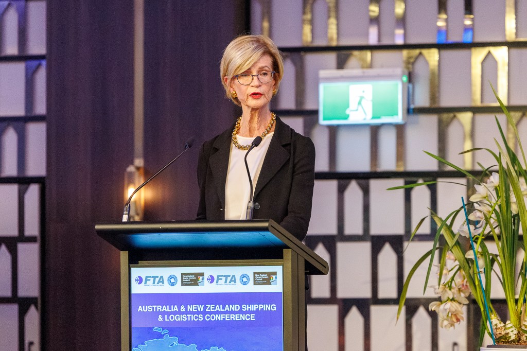 A blonde woman wearing a dark suit coat speaking from behind a podium that reads "Australia and New Zealand Shipping and Logistics Conference There is a plant to her left and an exit sign behind her.