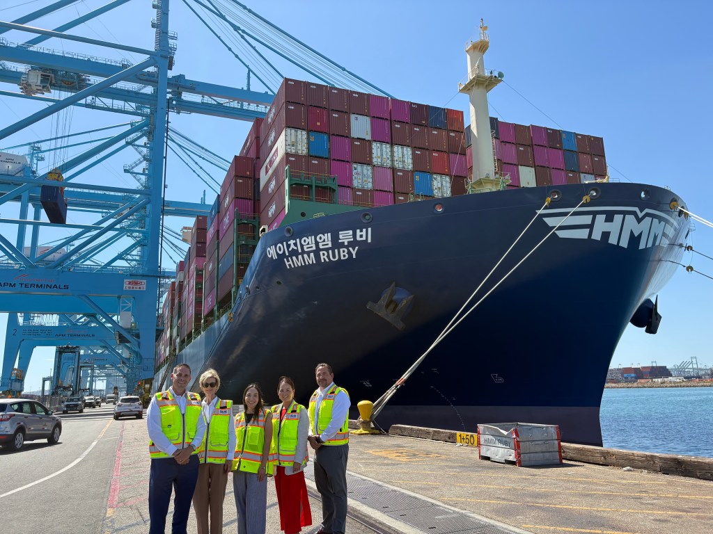 At a shipping pier with gantry cranes and a shipping vessel in the background, a group of 5 people in bright yellow safety vests pose for a photo.