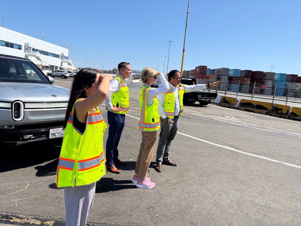 On pavement with shipping containers in the background, a group of people in bright safety vests looks in the direction to which a man from the group is pointing.
